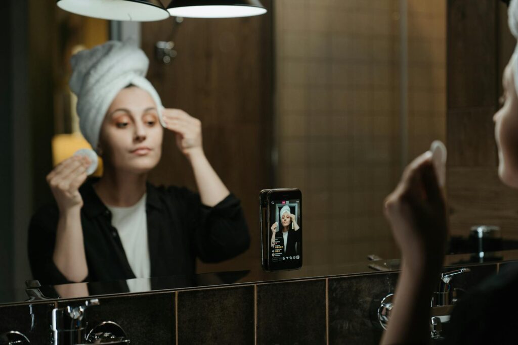 A woman applies skincare products while filming herself in a bathroom mirror with a smartphone.