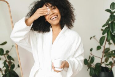 Happy woman in bathrobe applying skincare routine with cotton pad indoors.